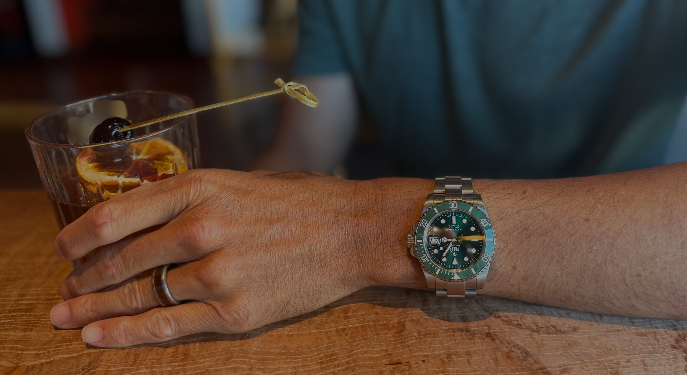 A person wearing an Ali'i Watch Company Waterman watch holds a Manhattan cocktail