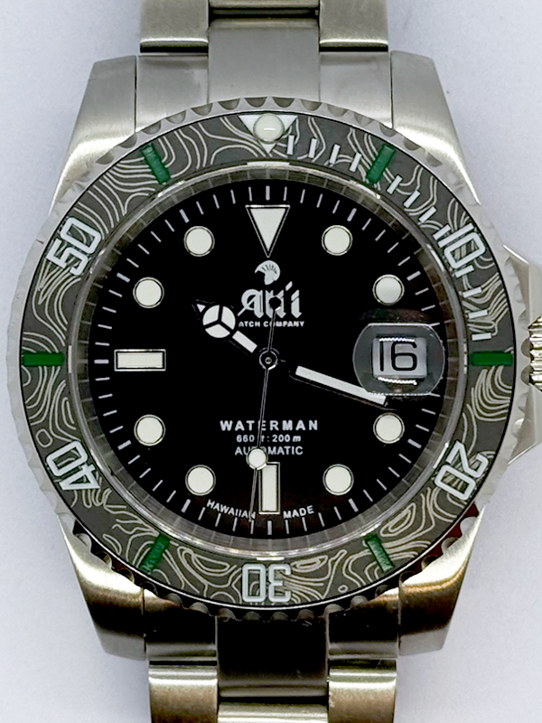 A close up of the dial on the Ali'i Watch Company Waterman watch with black dial and black carbon fiber bezel with green minute markers.